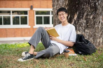 A positive, nerd Asian male student is writing some ideas in a book under the tree in a campus park.