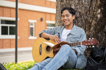Young positive Asian male is resting under the tree with his guitar, practicing playing guitar.