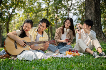 A lovely Asian couple is playing a guitar together while enjoying a picnic with their friends.