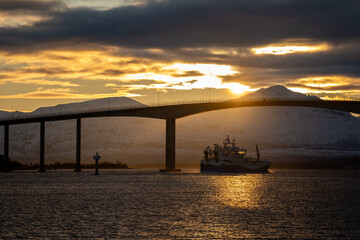 Statek przepływający pod wysokim mostem, na tle zachodu słońca i gór w Norwegii, Tromso