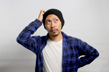 Puzzled young Asian man, dressed in a beanie hat and casual shirt, is depicted in a moment of contemplation, scratching his head, pondering a solution while standing against white background.
