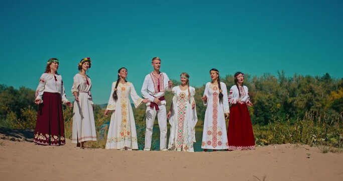 Group of talented people in national Slavic gowns singing songs on river shore in summer, 4K, Prores
