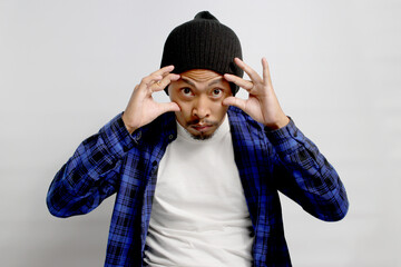 Sleepy, tired young Asian man in a beanie hat and casual outfit is struggling to open his eyes, displaying signs of morning fatigue or exhaustion, isolated on white background.