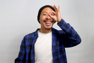 Excited young Asian man, wearing a beanie hat and casual shirt, is making an OK gesture with his...