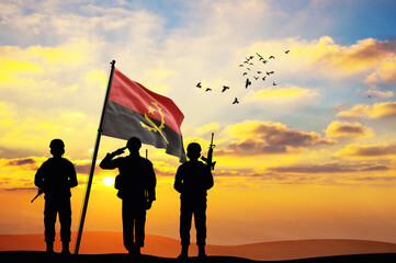 Silhouettes of soldiers with the Angola flag stand against the background of a sunset or sunrise....