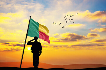 Silhouette of a soldier with the Benin flag stands against the background of a sunset or sunrise....