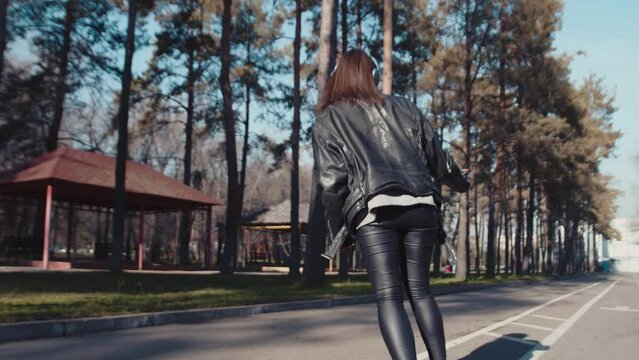 Back view of a stylish young girl in headphones and leather clothes riding on a skateboard on a path in a city park on a sunny day. Concept of interesting autumn urban leisure