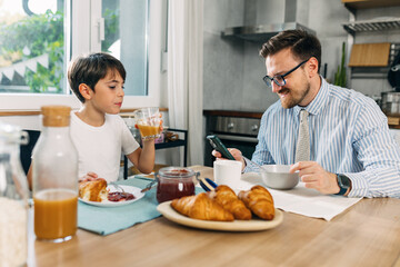 father is using mobile phone and having breakfast with his teenage son.