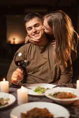 Happy young couple in love drinking wine, wife hugging and kissing her husband, celebrating Valentine's day having romantic dinner with candles at home