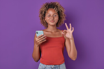 Naklejka premium Young thoughtful attractive African American woman zoomer touching face looks to side and holds mobile phone choosing words for important sms message in messenger app stands posing in purple studio.