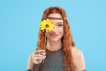 Beautiful young hippie woman covering eye with sunflower on light blue background