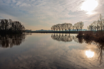 Landscape photo with a large lake and bare trees as silhouettes on the shore. It is a windless day and the water surface is very smooth. The low sun is just breaking through the cloudy sky.