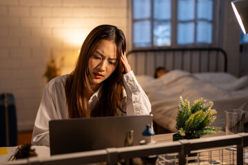 Depressed Asian young woman overworking while husband sleeping at night. 