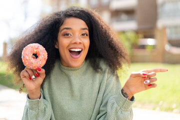Young African American woman holding a donut at outdoors surprised and pointing finger to the side