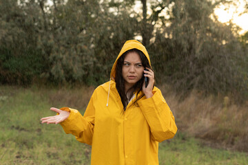 Upset girl in a yellow raincoat under heavy rain in the park calls a taxi using a smartphone