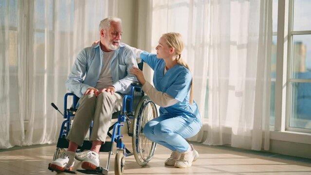 Nurse talking senior man in wheelchair. Female social welfare physical therapist looks after senior male, listening, providing medical care at home. Living with legs paralysis, special needs people.