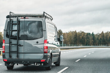 Fototapeta na wymiar Camper van on the public road. Offroad modern van for traveling with a ladder on the back and an additional baggage roof rack on the top. Concept of road journey and adventures.