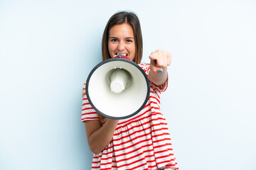 Young caucasian woman isolated on blue background shouting through a megaphone to announce something while pointing to the front