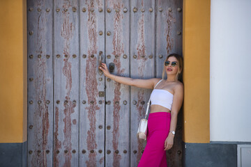 South American woman, young, beautiful and blonde with white top, pink pants and sunglasses, posing leaning on the frame of a wooden door. Trend concept, beauty, fashion.