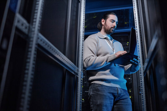 Young IT professional using laptop and standing near metal frame at server room