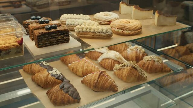 Assorted sweet baked goods on display shelves in bakery. There are croissants, eclairs, cakes and trifles for sale