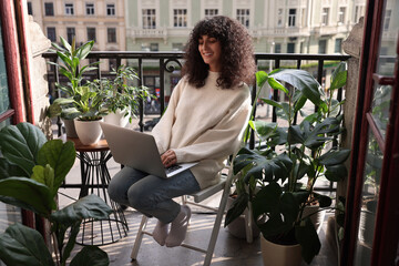 Beautiful young woman using laptop surrounded by green houseplants on balcony