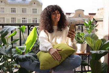 Beautiful young woman using smartphone surrounded by houseplants on balcony