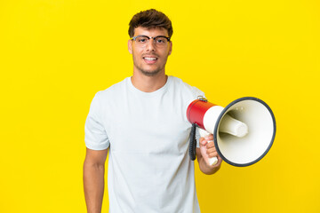 Young caucasian handsome man isolated on yellow background holding a megaphone and smiling a lot