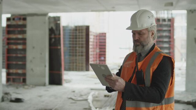 Medium shot of middle-aged Caucasian bearded contractor in safety vest and hard hat taking notes and photos on digital tablet while checking work at unfinished construction site