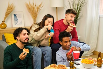 Group of diverse friends eating pizza and playing video games in shared apartment.
