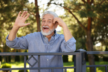 Neighbor greeting. Happy senior man waving near fence outdoors