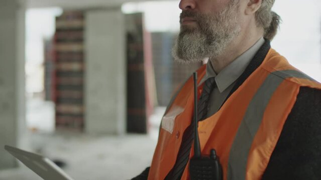 Tilting shot of confident adult Caucasian foreman with grey hair and beard in safety uniform using digital tablet at construction site while checking work of builders