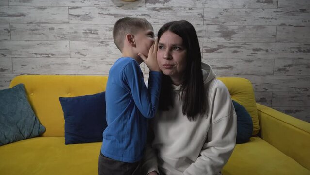 Medium shot. Cute son tells his mother a secret, whispers in her ear in the room, mom is surprised by what she heard. they spend their free time at home together