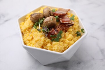 Cooked cornmeal with bacon, mushrooms and microgreens in bowl on white marble table, closeup