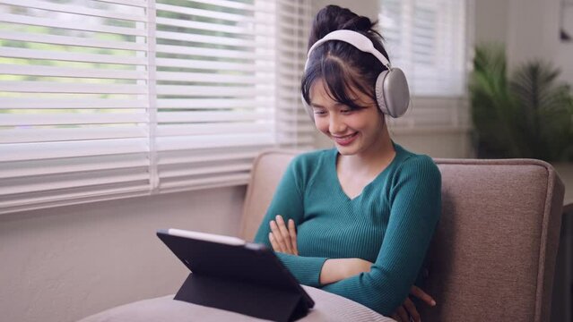 An Asian young woman enjoys a comedy show on her laptop, wearing headphones while seated on her home sofa during the weekend. Capture the joy of relaxation and entertainment
