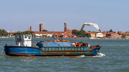 Fototapeta na wymiar A seagull flies over an old ship with a crane in the lagoon of Venice, Italy, with the island of Murano in the background