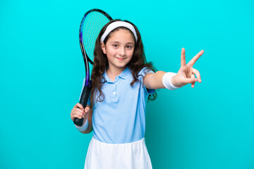 Little caucasian girl playing tennis isolated on blue background smiling and showing victory sign