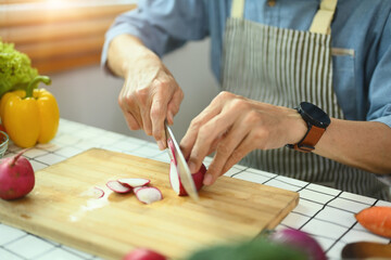 Senior man wearing apron cooking healthy food at home, slicing fresh radishes on cutting board. - obrazy, fototapety, plakaty