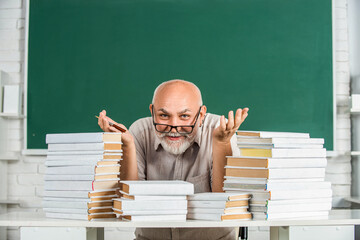 Teacher with books in school. Man elementary school teacher standing in classroom. Old man teachers on green board. Portrait male teacher in classroom. Funny teacher hold many books at school