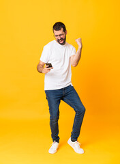 Fototapeta na wymiar Full-length shot of man with beard over isolated yellow background with phone in victory position