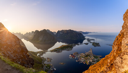 Midnight sun casts a warm glow over the panoramic view from Reinebringen, with the sharp peaks of the Lofoten Islands enveloping the calm waters of the fjord, and village of Reine nestled below