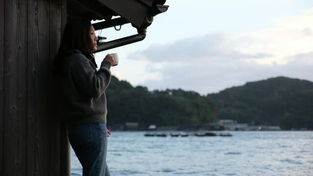 A woman standing and drinking coffee by the ocean in Ine village, Japan