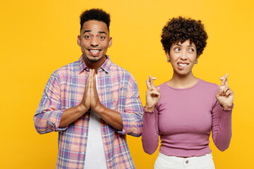 Young couple two friend family man woman of African American ethnicity wear purple casual clothes together hold hands folded in prayer gesture keep fingers crossed isolated on plain yellow background