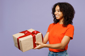 Side view little kid teen girl of African American ethnicity wear orange t-shirt hold in hand present box with gift ribbon bow isolated on plain pastel purple background. Childhood lifestyle concept.