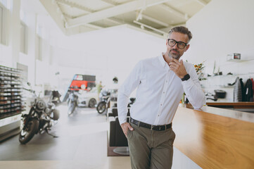 Male pensive successful businessman man wear classic shirt prop up chin look camera standing at office table in dealership store indoors want to buy new motorbike or car. Business lifestyle concept. - obrazy, fototapety, plakaty
