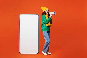 Full body side view young woman wear green knitted sweater yellow hat scarf big huge blank screen mobile cell phone smartphone with area scream in megaphone isolated on plain orange red background.