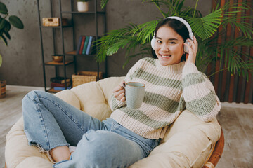 Obraz premium Sideways young woman wears casual clothes listen to music in headphones drink tea sits on armchair stay at home hotel flat rest relax spend free spare time in grey living room indoor. Lounge concept.