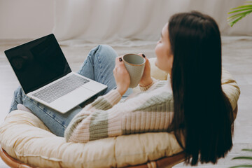 Young IT woman wears casual clothes sits on armchair work use blank screen laptop pc computer drink coffee stay at home hotel flat rest relax spend free spare time in grey living room. Lounge concept. - obrazy, fototapety, plakaty