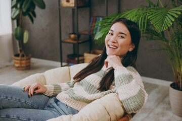 Sideways young woman of Asian ethnicity she wear casual clothes looking camera sits on armchair stay at home hotel flat rest relax spend free spare time in living room indoor. People lounge concept.