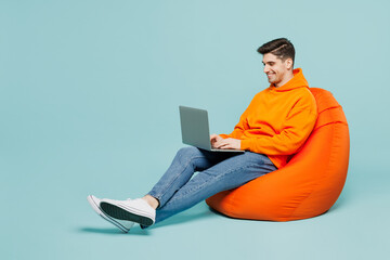 Full body fun young IT man he wear orange hoody casual clothes sit in bag chair hold use work on...